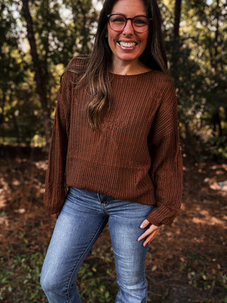Woman wearing a brown sweater and blue jeans standing in a forest.- ava wray boutique