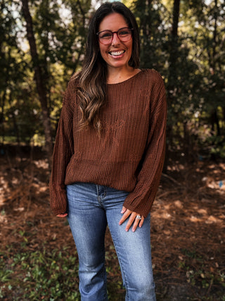 Woman wearing a brown sweater and blue jeans standing in a forest- ava wray boutique